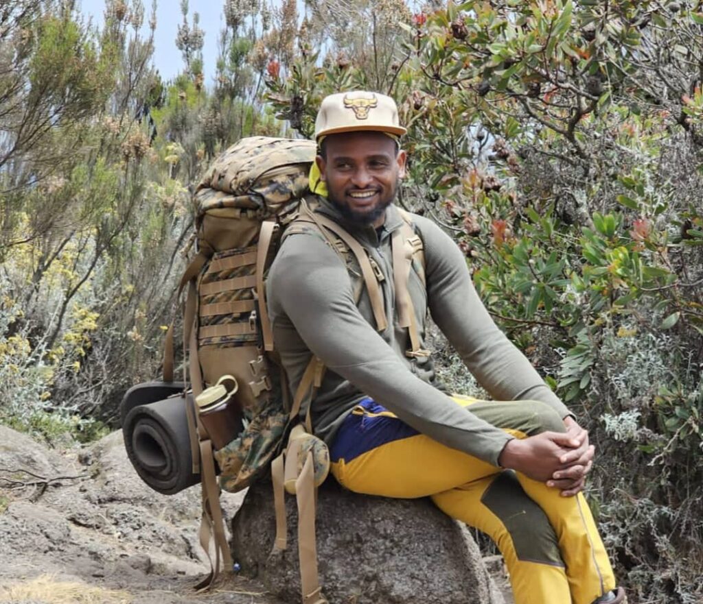 A smiling man sits on a rock while hiking, wearing a beige cap, long-sleeve shirt, and yellow pants. He has a large backpack and is surrounded by green foliage and shrubs.