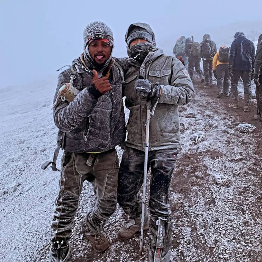 Two people in winter gear pose on a snowy path, surrounded by others hiking in cold weather. One person gives a thumbs up, while the other holds a walking stick. The ground is covered with snow, and the sky is foggy.