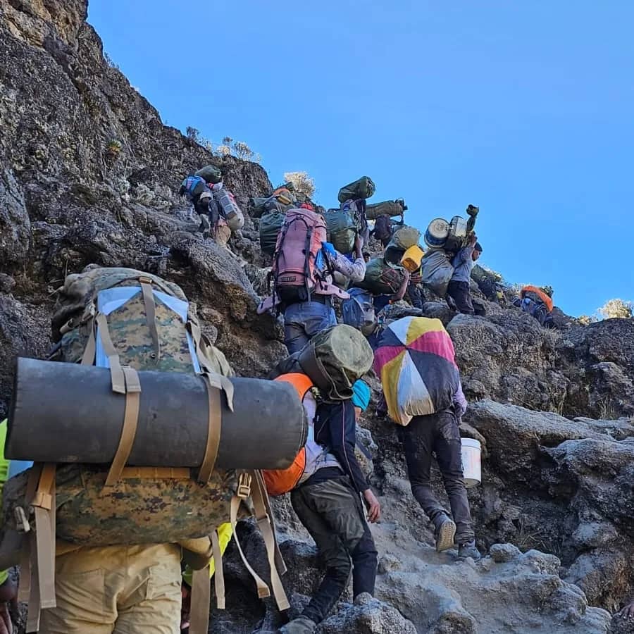 A line of hikers with large backpacks ascend a rocky mountain trail under a clear blue sky. The group appears to be on a challenging climb, surrounded by rugged terrain and sparse vegetation.