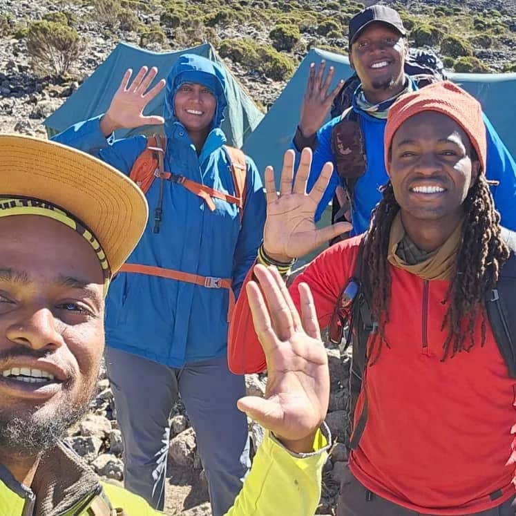 Four people in outdoor gear smile and raise their hands in a greeting gesture, with tents and greenery in the background.