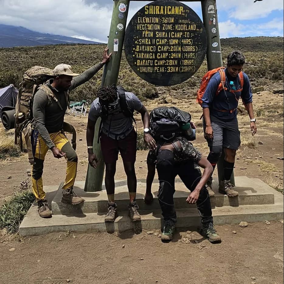 Four hikers rest at a signpost for Shira Camp on Mount Kilimanjaro, with backpacks and outdoor gear. They appear tired but cheerful, standing or crouching near the sign in a rocky and grassy landscape.