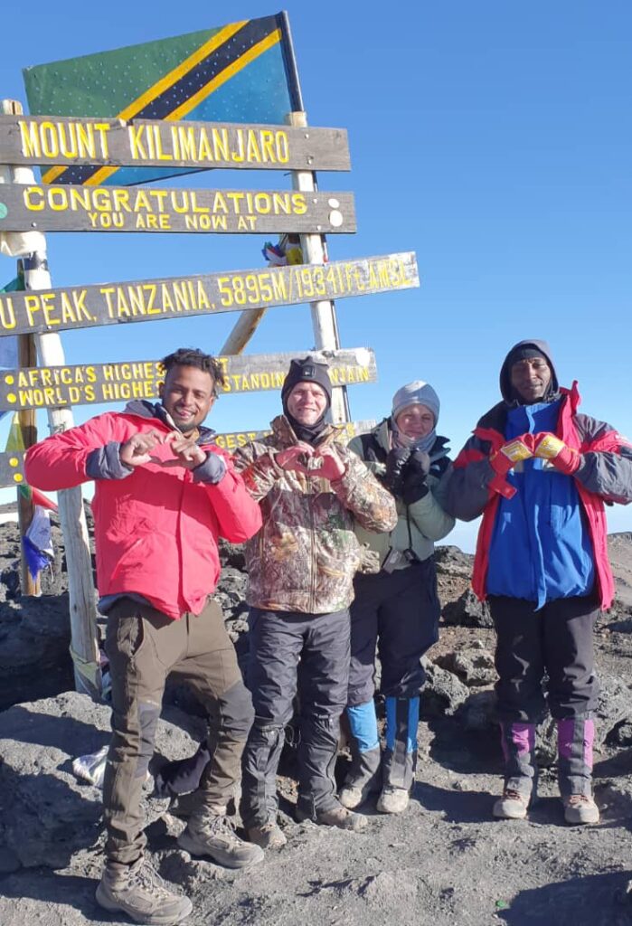 Four people smile and gesture heart signs with their hands at the summit of Mount Kilimanjaro. They stand in front of a sign marking the peaks height. The sky is clear, and they are dressed in warm hiking gear.