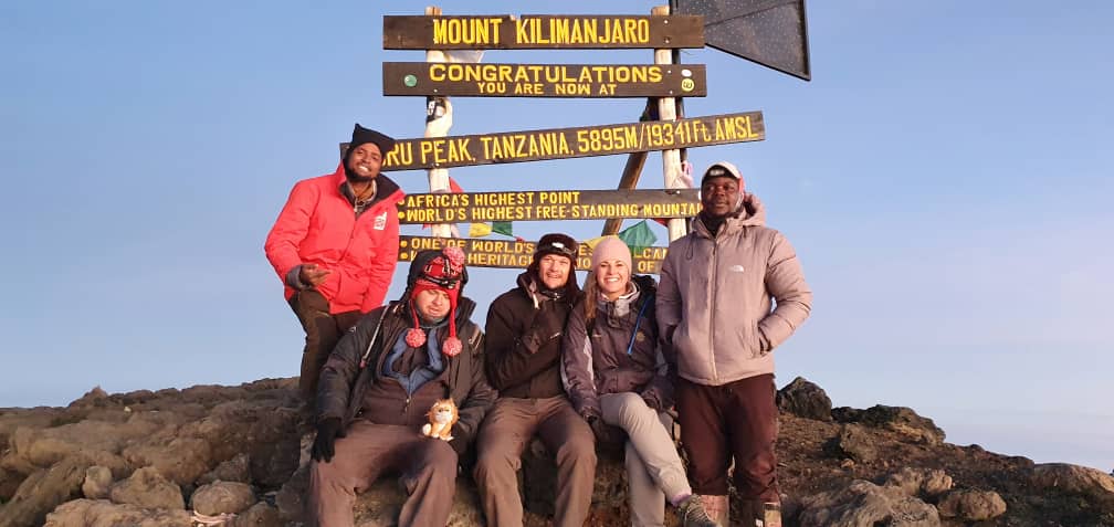 Five people pose at the summit of Mount Kilimanjaro, standing and sitting in front of the sign indicating Uhuru Peak's elevation and status.