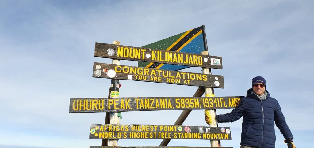 
	
A person in a winter jacket and sunglasses stands beside a sign on Mount Kilimanjaro. The sign reads Congratulations, you are at Uhuru Peak, Tanzania, 5895m/19341 ft AMSL, Africas highest point, worlds highest free-standing mountain.